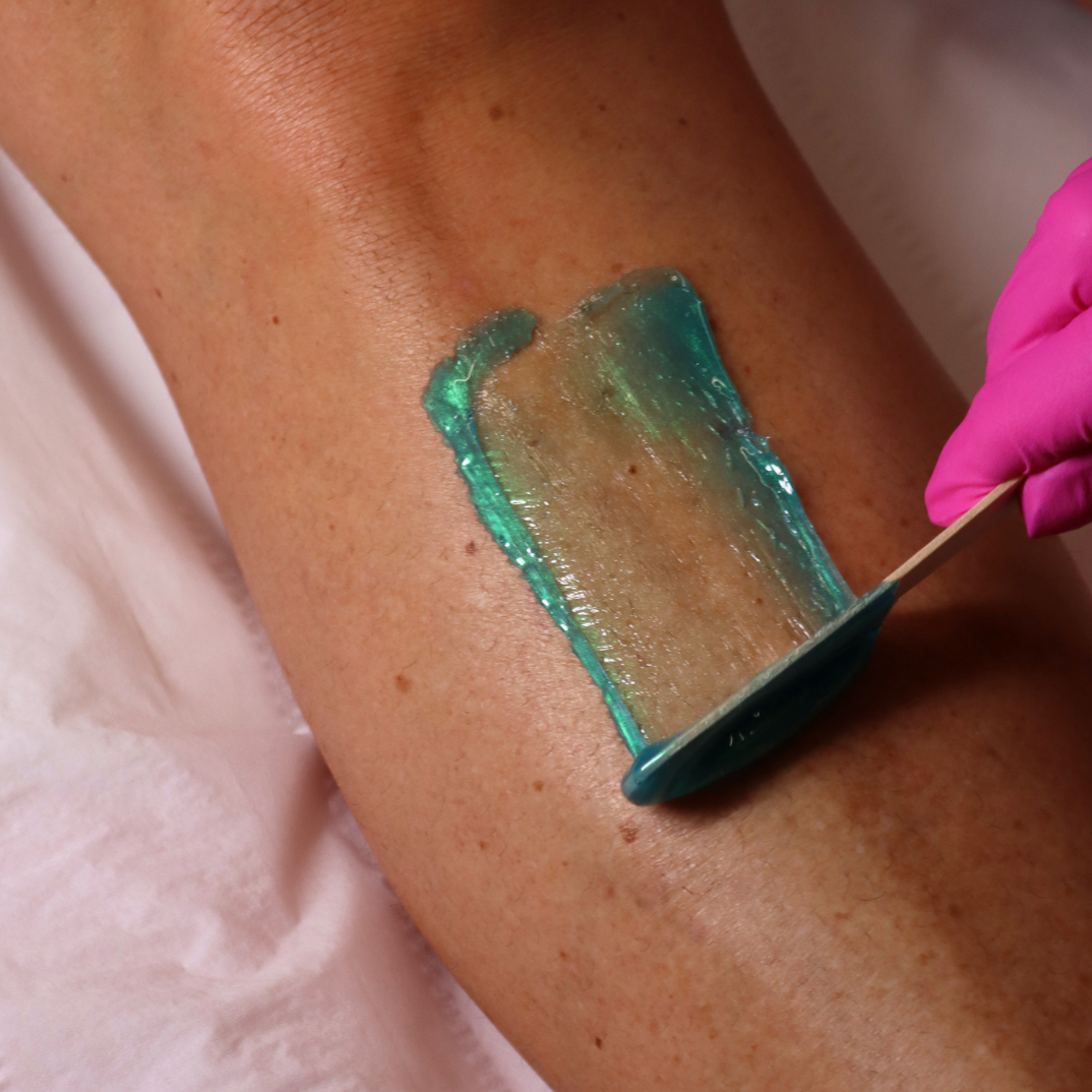 A close-up of a leg being treated with a teal hair removal wax, held by a hand wearing a pink glove and spread with a wooden spatula.